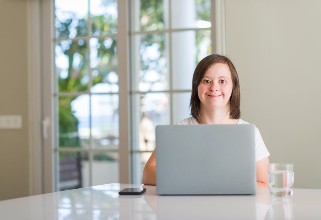 Down syndrome woman at home using computer laptop exploring customised employment and discovery with a happy face standing and smiling with a confident smile showing teeth